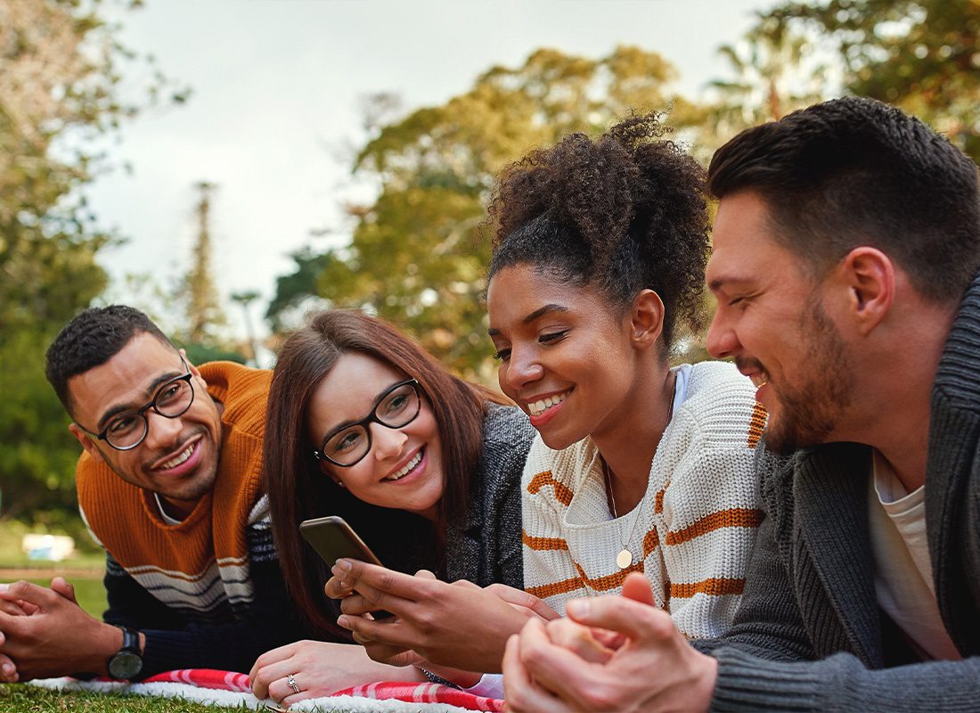 Read Our Reviews - A Group of Friends Smiling Together Because of Something on a Smartphone at the Park on a Nice Day